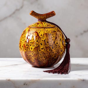 May include: A decorative ceramic jar with a lid and a tassel. The jar is a mix of yellow and brown with a textured pattern. The lid has a branch-like handle. A maroon tassel hangs from the jar, adding a decorative element.