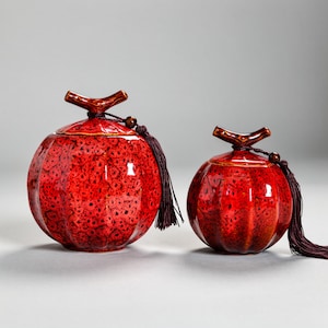 May include: Two red ceramic jars with lids, each featuring a textured surface and a dark brown tassel. The jars have a round shape with a stem-like handle on top. The jars are a deep red color with darker spots.