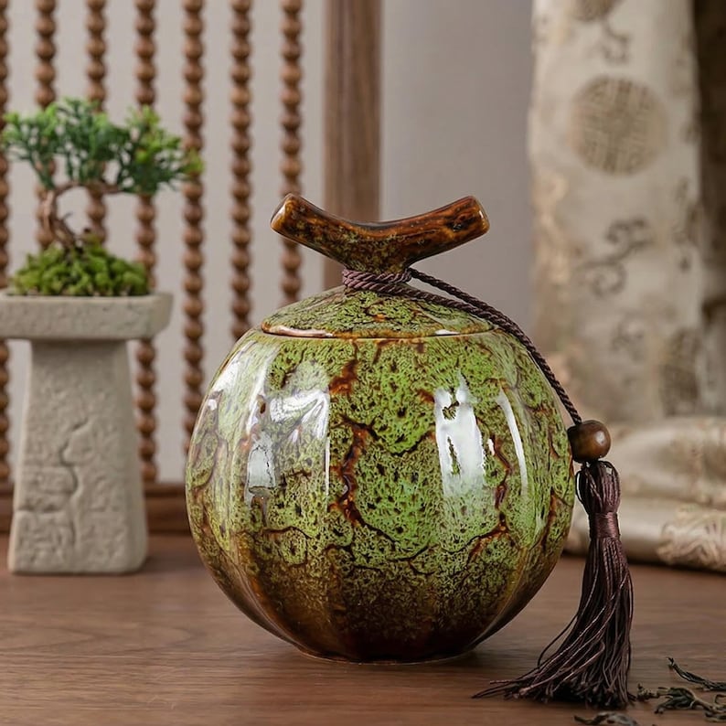 May include: A green and brown ceramic jar with a lid and a decorative tassel. The jar has a unique, textured pattern and a wooden handle on top. The jar is round and sits on a wooden surface.
