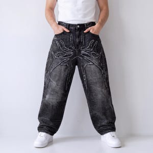May include: Black denim jeans with a unique, stylized design on the front. The jeans have a faded, distressed look and a relaxed fit. The model is wearing white sneakers and a white tank top.