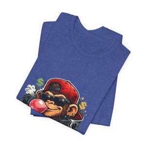 May include: Blue t-shirt featuring a cartoon monkey wearing a red cap, sunglasses, and a gold earring, blowing a pink bubble. The design includes dollar signs and smoke clouds.