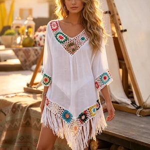 May include: White beach cover-up with a V-neck and crochet detailing. The cover-up features colorful crochet accents around the neckline, sleeves, and hem, with a fringe trim. The sleeves are three-quarter length.