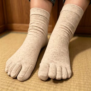 May include: Beige toe socks, with each toe individually separated. The socks are made from a soft, textured fabric and feature ribbed cuffs. The feet rest on a woven mat, with a wooden wall visible in the background.
