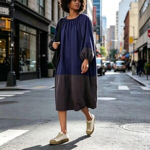 May include: A navy blue and gray color-block dress with long sleeves and a round neckline. The dress is knee-length and paired with off-white sneakers with a brown sole. The dress is worn in an urban setting.