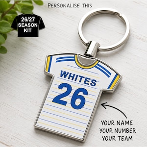 May include: A metal keyring shaped like a sports jersey with a white background, thin blue and yellow horizontal stripes, the text WHITES, and the number 26 in blue. The keyring includes a silver split ring for keys and a metallic border.