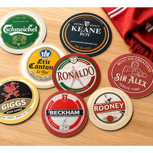 May include: A selection of circular drinks coasters featuring football-themed designs with names like "Schmeichel", "Keane Roy", "Eric Canton", "Ronaldo", "Giggs", "Beckham", "Sir Alex", and "Rooney". The coasters are on a wooden table, with a pint of beer and a red scarf.