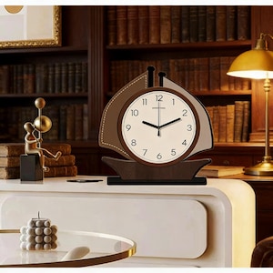 Puede incluir: Un reloj de escritorio con forma de velero, marrón y crema. La esfera del reloj tiene números y manecillas negras. El reloj está sobre una base marrón oscuro. El fondo incluye una estantería, una lámpara y objetos decorativos.