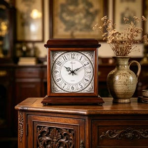 May include: A square, wooden mantel clock with a white face and black Roman numerals. The clock has black hands and a dark brown wooden frame. The clock sits on a dark wooden cabinet, with a vase of dried flowers to the right.