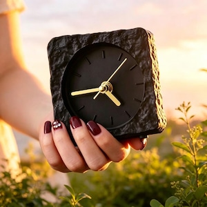 Könnte beinhalten: Eine quadratische, schwarze Analoguhr mit goldenen Zeigern und einem schwarzen Zifferblatt. Die Uhr hat einen strukturierten, steinartigen Rahmen. Die Uhr wird in einer Hand mit dunkelrotem Nagellack gehalten. Der Hintergrund ist verschwommen mit warmem, goldenem Licht.