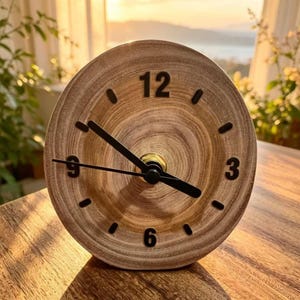 Könnte beinhalten: Eine runde Holzuhr mit hellbraunem Finish. Das Zifferblatt hat schwarze Zahlen an den Positionen 12, 3, 6 und 9 Uhr, mit schwarzen Zeigern und einer goldfarbenen Mitte. Die Uhr steht auf einer Holzoberfläche.