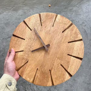 Könnte beinhalten: Eine kreisförmige Wanduhr aus hellbraunem Holz mit sichtbarer natürlicher Maserung. Das Zifferblatt verfügt über zwölf gleichmäßig verteilte radiale Einkerbungen als Stundenmarkierungen und zwei dunkelbraune Holzzeiger in der Mitte.