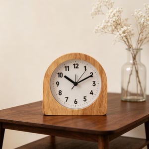 May include: An arched tabletop clock with a light wood grain frame and a white circular face. The clock features black Arabic numerals from 1 to 12, black hour and minute hands, and a thin black second hand, all set against a clean white background.