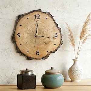 Puede incluir: Un reloj de pared de madera hecho de una sección transversal de un tronco de árbol. La esfera del reloj presenta números negros en las posiciones 3, 6, 9 y 12. Las manecillas del reloj son doradas. El reloj está montado en una pared blanca. Objetos decorativos sobre una superficie de madera.