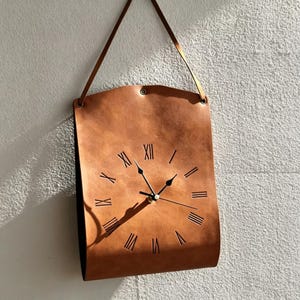 May include: A tan leather wall clock with a curved rectangular shape, featuring black Roman numerals cut into the surface and black clock hands. The clock is suspended by a matching tan leather strap attached to the top corners with metal rivets.