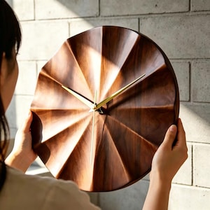 Könnte beinhalten: Eine Wanduhr aus Holz mit einem einzigartigen, geometrischen Design. Das Zifferblatt hat eine warme braune Farbe mit einem radialen Muster. Goldfarbene Zeiger zeigen die Uhrzeit an. Die Uhr wird vor einer hellen Backsteinmauer gehalten.