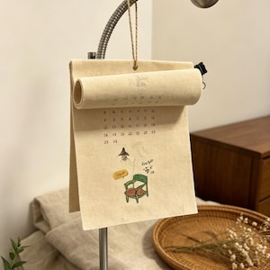 May include: A cream-colored fabric calendar hanging from a silver metal lamp. The calendar features a printed grid of dates, a small illustration of a green chair, a lamp, and text. The top portion of the fabric is rolled up and secured with a black clip.