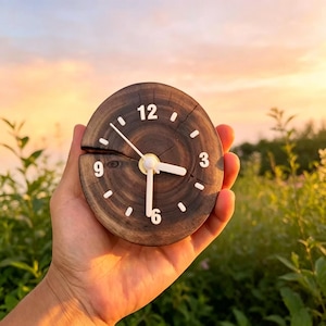 Pode incluir: Um pequeno relógio de madeira redondo com algarismos e ponteiros brancos. O mostrador do relógio é feito a partir de uma seção transversal de um tronco de árvore, mostrando o grão natural da madeira. O relógio é segurado em uma mão contra um fundo de pôr do sol.