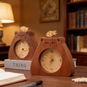 May include: Two wooden cat-shaped clocks with a fish on top. The clocks have a light-colored clock face with gold-colored hands. The word "THING" is visible on a book beneath one of the clocks. The clocks are brown.