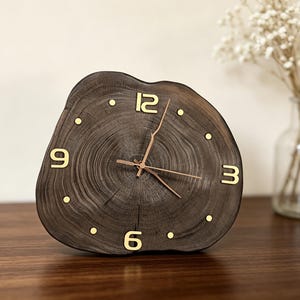 May include: A wall clock crafted from a dark brown natural wood slice with visible tree rings. The clock face features gold-coloured numerals at the 3, 6, 9, and 12 o'clock positions, with gold dots for the remaining hours and thin gold hands.
