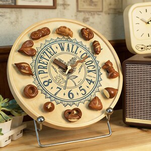 Puede incluir: Un reloj de pared redondo de madera con marcadores en forma de pretzel. La esfera del reloj tiene un diseño azul y blanco con las palabras "Pretzel Wall Clock". El reloj está sostenido por un soporte de metal plateado.