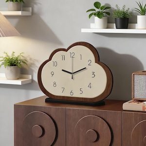 May include: Cloud-shaped wooden clock with a cream-coloured face and black numerals. The clock has a dark brown wooden frame and a black base. The clock is sitting on a dark brown wooden cabinet. The clock measures approximately 25 cm in height.