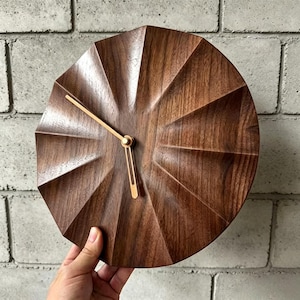Könnte beinhalten: Eine runde Holzuhr mit einem geometrischen Sternmuster. Die Uhr ist aus dunkelbraunem Holz gefertigt und hat goldfarbene Zeiger. Die Uhr wird in einer Hand vor einer weißen Backsteinmauer gehalten.