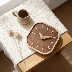 May include: A square wooden clock with a walnut face and light wood frame. White markers indicate the hours. A small beige pitcher and two dried flower stems sit nearby. The clock is on a wooden surface.