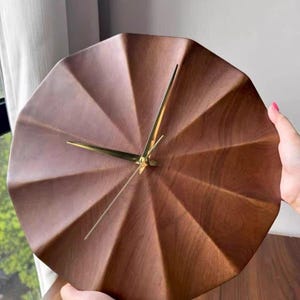 Op de afbeelding: Een ronde wandklok van donkerbruin hout met een gefacetteerd, geplooid oppervlak. De klok heeft drie dunne, gepolijste goudkleurige metalen wijzers in het midden van de wijzerplaat, zonder zichtbare cijfers of uurmarkeringen op het hout.