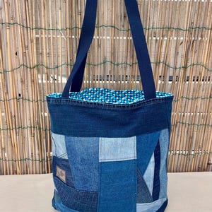 Puede incluir: Un bolso tote de mezclilla patchwork con correas azul marino. La bolsa está hecha de varios tonos de mezclilla azul, creando un patrón geométrico texturizado. El forro interior presenta un estampado de lunares azules y blancos. La bolsa está diseñada para transportar objetos.