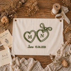 May include: Cream-colored fabric banner with embroidered green hearts, initials "M" and "N", and the date "June 15.06.2026". The banner is surrounded by dried flowers, lace, and stationery, suggesting a wedding theme.
