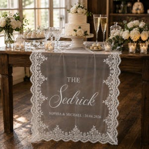 Puede incluir: Una mesa de recepción de bodas con una tarta blanca de dos pisos, copas de champán y arreglos florales. Un camino de mesa transparente con bordes de encaje y el texto "THE Sedrick" está colocado sobre la mesa.