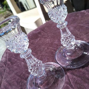 May include: Two clear glass candle holders with diamond-cut patterns. The candle holders have a flared top, a decorative stem, and a round base. The glass reflects light, creating a sparkling effect. These are suitable for home decor.