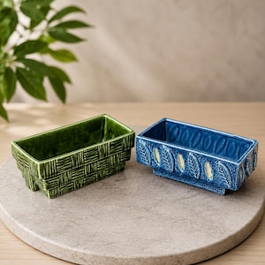 May include: Two rectangular ceramic planters. The green planter features a textured basket-weave pattern on its exterior. The blue planter displays a repeating embossed leaf motif along its sides. Both containers have a glossy finish and a raised base.