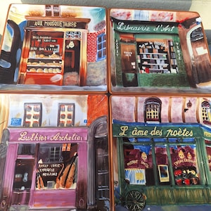 May include: Set of four square ceramic plates featuring colorful illustrations of Parisian storefronts. Each plate depicts a different shop: a bakery, bookstore, violin shop, and a cafe. The plates have a vintage, hand-painted aesthetic.