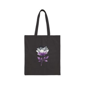 May include: Black tote bag featuring a watercolor flower design. The flower has petals in shades of gray, white, and purple, with a matching purple stem and leaves. The bag has two black handles.