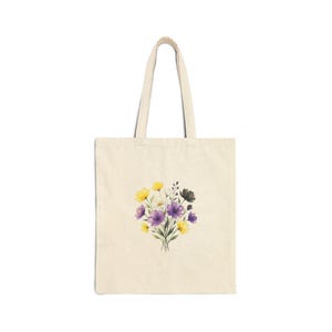 May include: A cream-coloured tote bag featuring a watercolour floral design. The bouquet includes yellow, purple, white, and black flowers. The bag has two handles and is made of a sturdy fabric, ideal for carrying items.