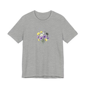 Op de afbeelding: Een grijze t-shirt met een aquarel bloemenontwerp. Het boeket bevat gele, witte, paarse en zwarte bloemen. Het shirt heeft korte mouwen en een ronde hals.