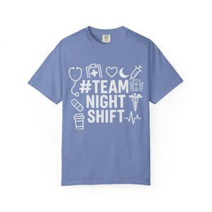 以下が含まれることがあります： 水色のTシャツで、白で「#TEAM NIGHT SHIFT」の文字と医療をテーマにしたグラフィックが描かれています。グラフィックには、聴診器、医療バッグ、心臓、月、注射器、病院、ピル、コーヒーカップ、心拍ラインが含まれています。