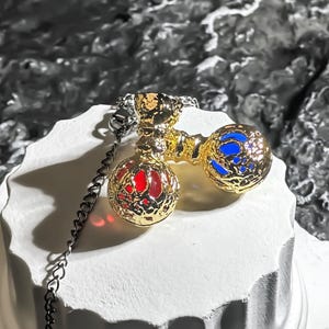 May include: A gold-colored pendant necklace featuring two orbs, one red and one blue, connected by a decorative gold piece. The orbs have a detailed, textured design. The necklace chain is silver-colored and hangs down.