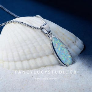 May include: A silver pendant necklace featuring a teardrop-shaped opal. The opal displays a mix of blue, green, and yellow hues. The necklace is presented on a seashell. Text reads "FancylucystudioGB" and "Handcrafted Jeweller".