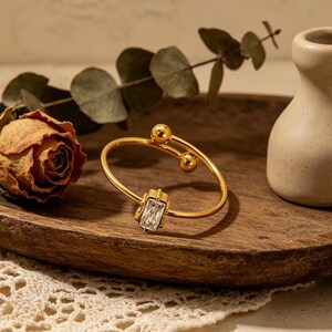 May include: A gold bracelet featuring a rectangular, clear gemstone. The bracelet has a distinctive design with two small gold spheres and a curved band. The jewellery is displayed on a wooden tray with a dried rose and greenery.