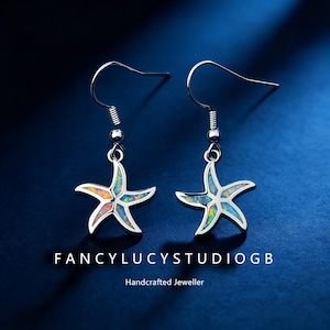 May include: Silver starfish earrings with colorful opal inlays. The earrings have a hook closure and are suspended from a small silver bead. The text "Fancylucystudiogb" and "Handcrafted Jeweller" are at the bottom of the image.