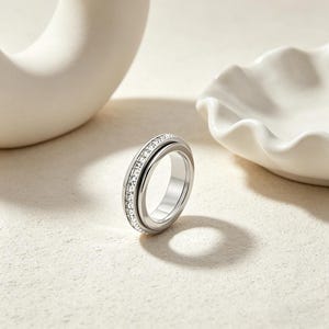 Peut inclure: Une bague argentée ornée d'une rangée de pierres claires et scintillantes. La bague est présentée sur un fond neutre, avec une sculpture blanche abstraite et une coupelle blanche festonnée. Le design de la bague est moderne et élégant.
