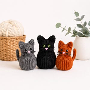 Puede incluir: Tres figuras de gato tejidas en gris, negro y naranja. Cada gato tiene una nariz rosa, bigotes negros y ojos verdes o negros. Los gatos están sentados. Una madeja de lana y una cesta están en el fondo.