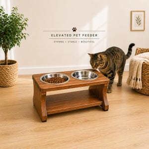 May include: A wooden elevated pet feeder with two stainless steel bowls. One bowl is filled with dry pet food. The feeder has a lower shelf and is labeled "ELEVATED PET FEEDER" with the words "STRONG • STABLE • BEAUTIFUL". A tabby cat is nearby.