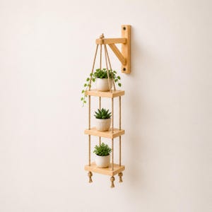 May include: A three-tiered hanging wooden shelf with jute rope supports. Each shelf holds a small potted plant. The top shelf is attached to a wooden bracket mounted on the wall. The plants have green leaves and are in white pots.