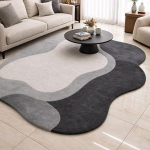 Op de afbeelding: Een modern vloerkleed met een abstract ontwerp. Het vloerkleed heeft een golvend patroon in grijstinten en wit, waardoor een zacht, getextureerd oppervlak ontstaat. Het vloerkleed staat in een woonkamer met een bank en salontafel.