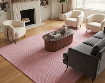 May include: A large rectangular area rug in a soft pink hue featuring a linear ribbed texture. The rug is positioned on a light-toned wooden floor in a living room setting, surrounded by three cream-colored armchairs and a dark gray sofa.