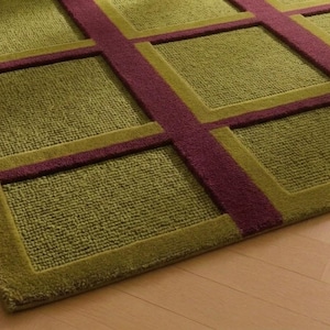 May include: A rectangular rug with a geometric pattern. The rug features a grid design with olive green squares and burgundy stripes. The rug is made of a soft, textured material and is placed on a light-colored wooden floor.