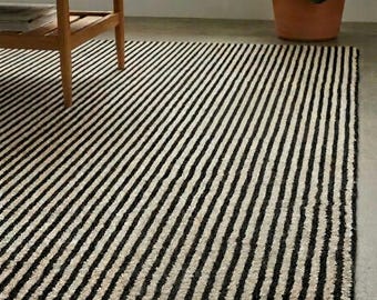 May include: A rectangular area rug featuring a repeating pattern of thin, vertical black stripes alternating with off-white, textured stripes. The rug lies flat on a smooth surface, extending from the foreground toward the back of the space.